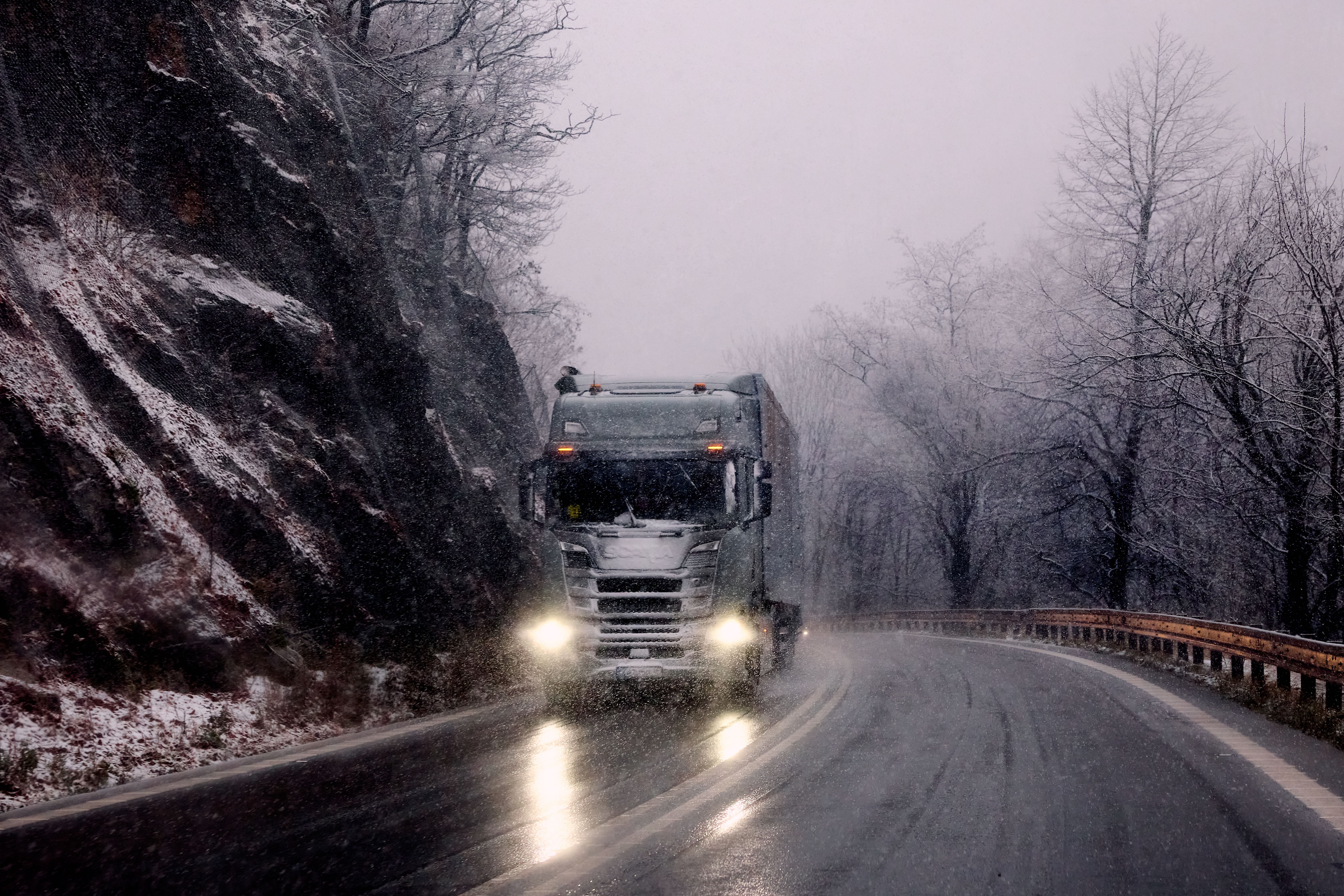 Semi truck driving through falling snow on a wet mountain highway with headlights on
